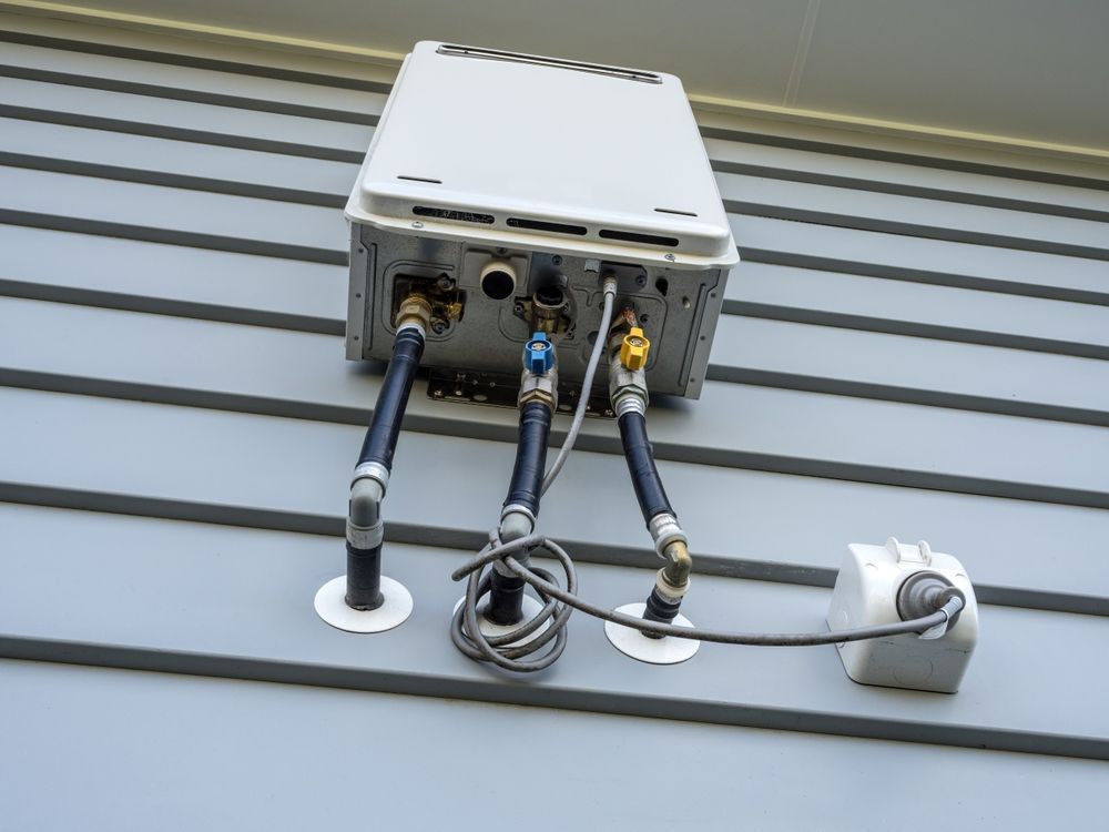 A white, outdoor tankless water heater is mounted on gray siding with black pipes. A small, white power supply is attached.