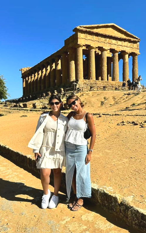 Two people pose in front of an ancient temple under a blue sky; they are smiling.