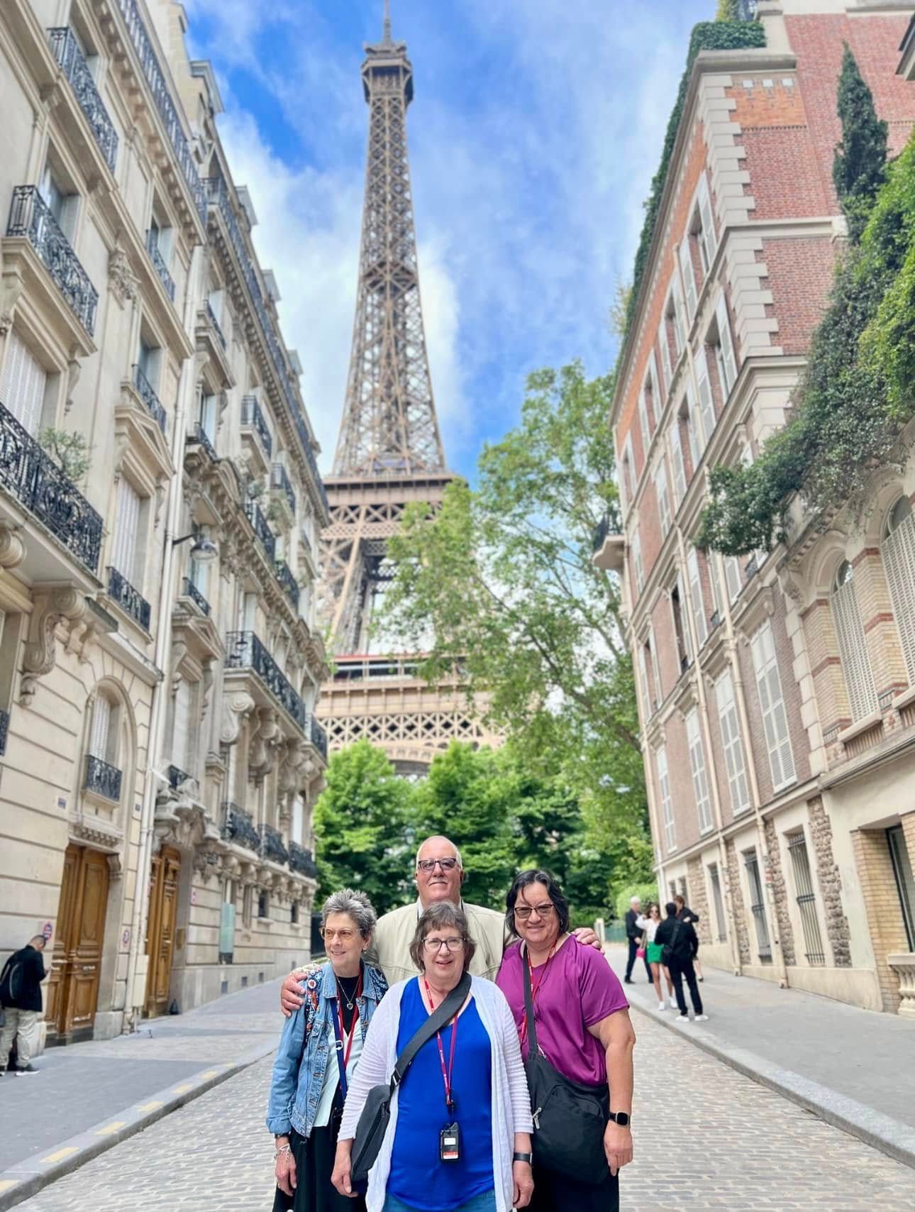 Four people pose in front of the Eiffel Tower, cobblestone street, tall buildings on each side.