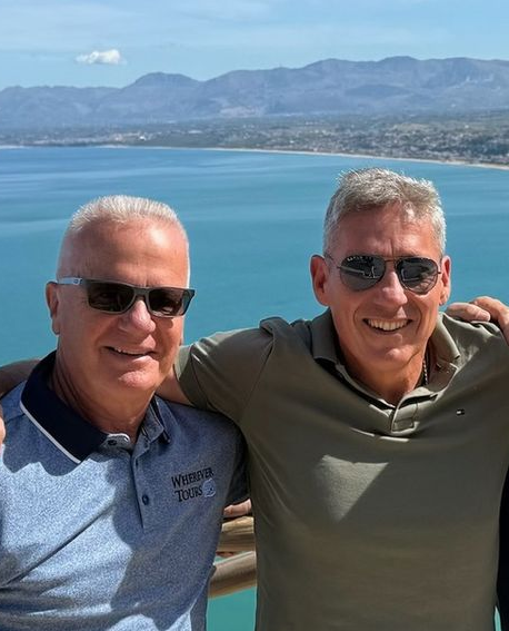 Two men in sunglasses smile at the camera, coastal scenery in background.
