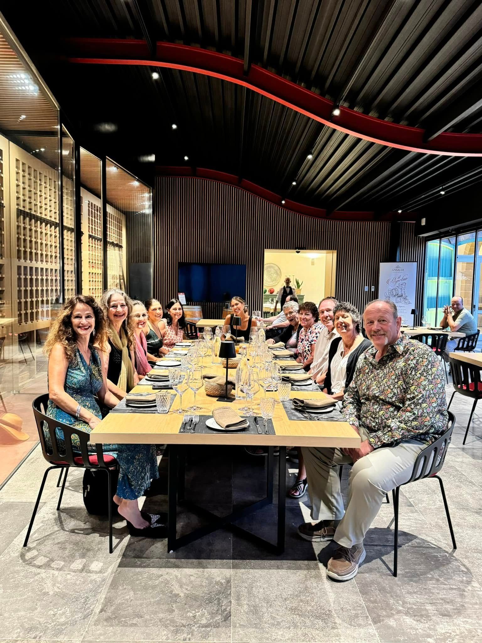 People seated at a long table in a restaurant. Dining room with decorative ceiling and wine display.