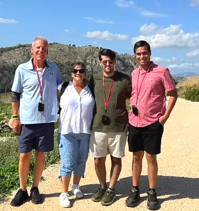 Four people pose outdoors on a sunny day. Mountain in the background. Two men, two women, all wearing casual clothes, smile.