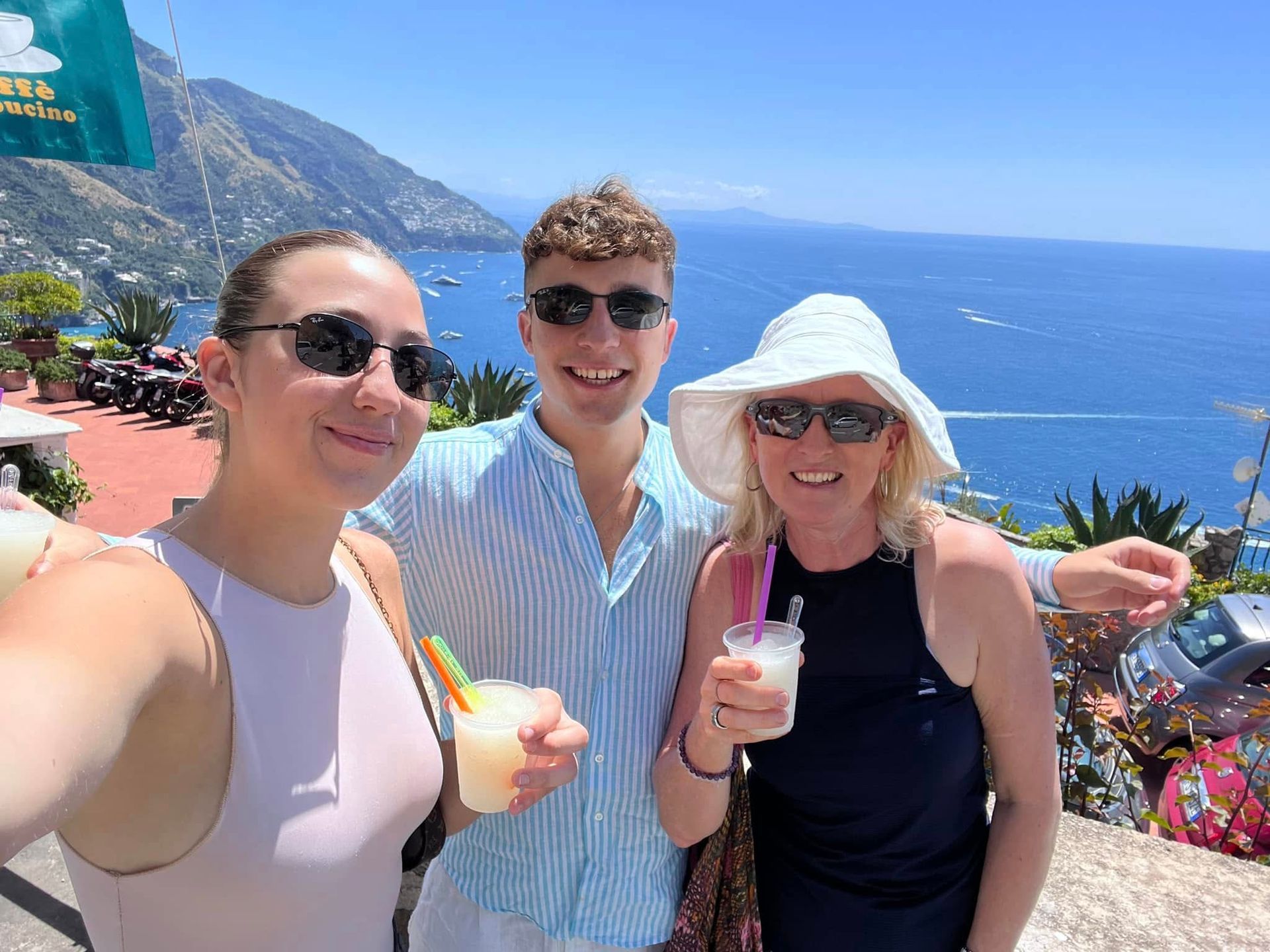 Three people with drinks smile at the camera, overlooking a coastal vista.