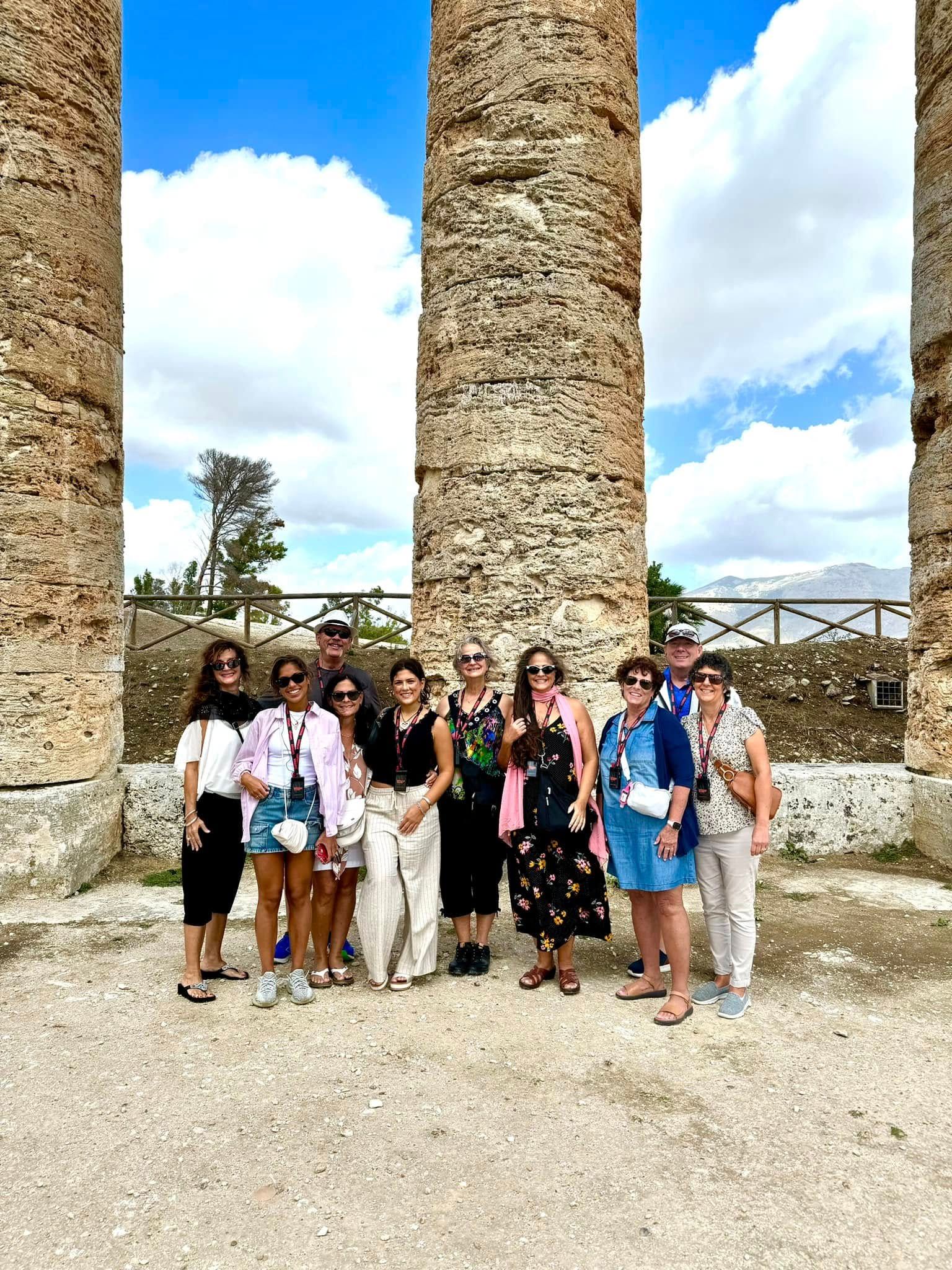 Group of people posing between ancient stone columns outdoors. Blue sky, sunny.