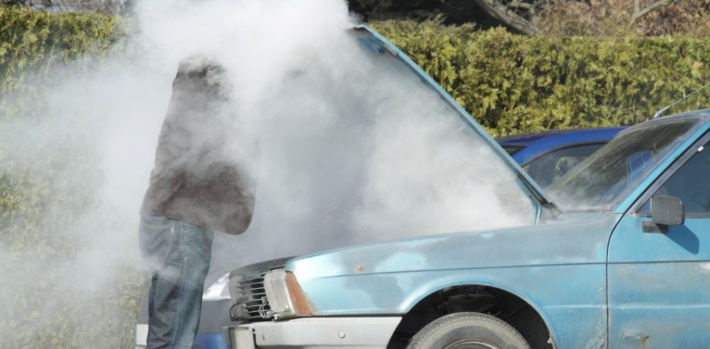 A man is standing next to a blue car with the hood open and steam coming out of it.