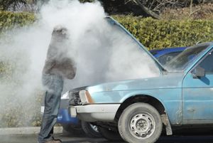 A man is standing next to a blue car with the hood open and smoke coming out of it.