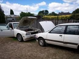 Two white cars are parked next to each other in a gravel lot.