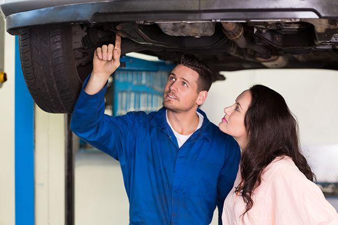 A man and a woman are looking under a car in a garage.