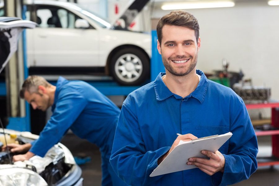 A mechanic is holding a tablet in a garage while another mechanic works on a car.