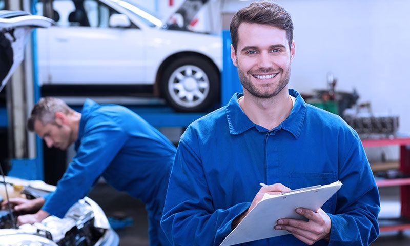 A mechanic is smiling while holding a clipboard in a garage.