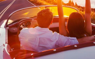 A man and a woman are sitting in a convertible car with their arms in the air.