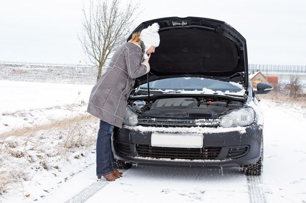 A woman is looking under the hood of a broken down car in the snow.