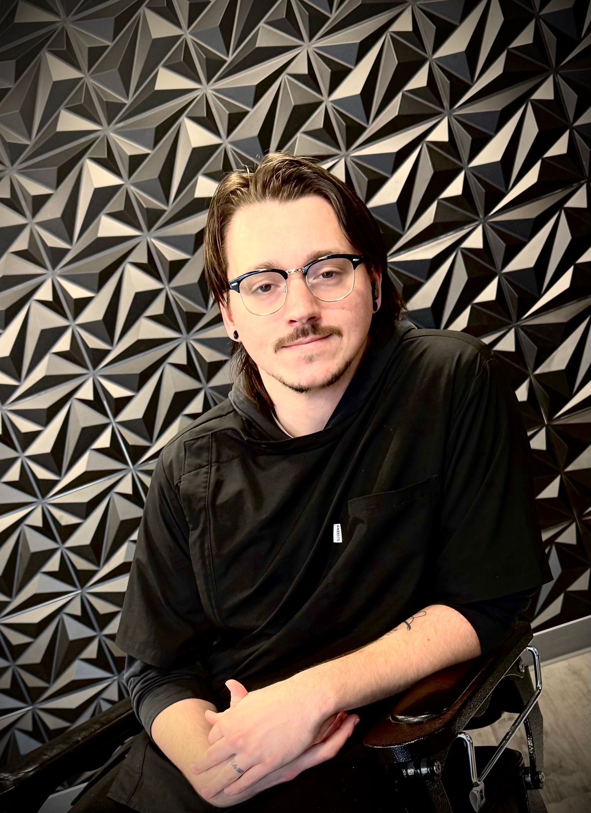 Man wearing glasses and black smock, sitting in front of a textured black and gray wall.