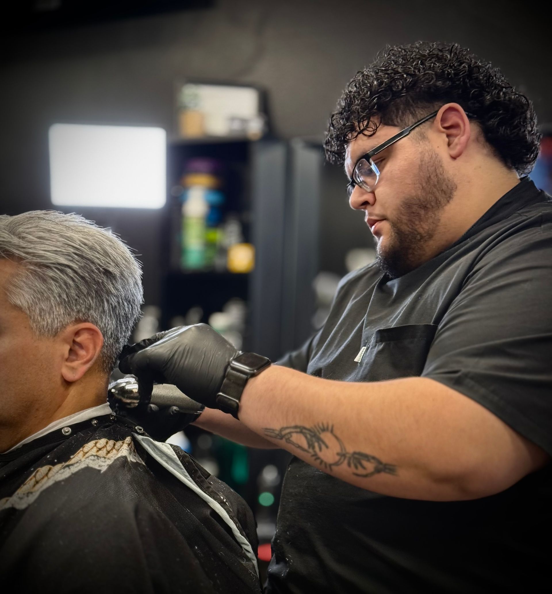 Barber cutting client's hair with clippers. Black gloves, tattoo on arm, in a barber shop.