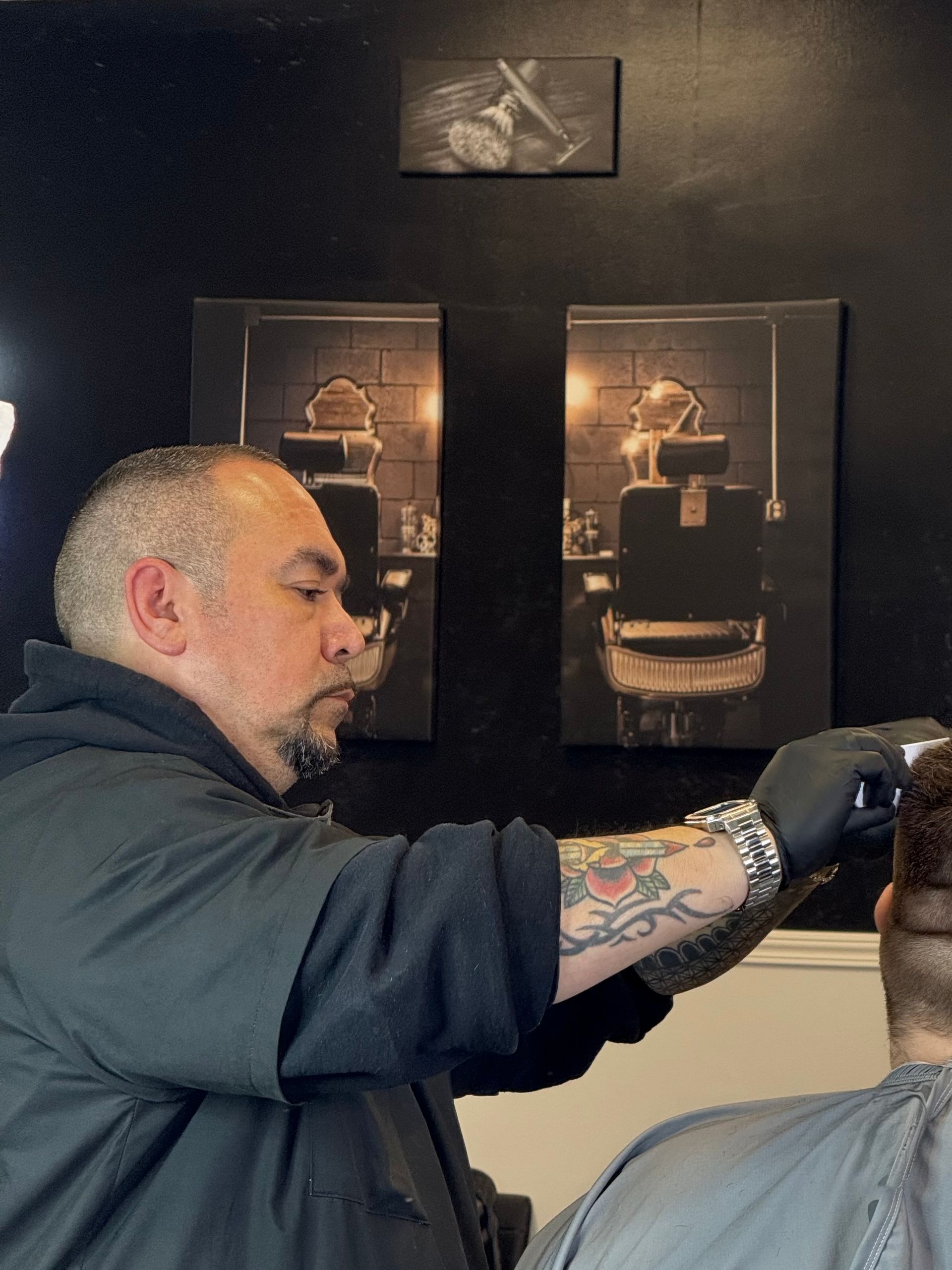 Barber giving a haircut to a client at a barbershop. He's wearing gloves and using clippers on the back of the client's head.