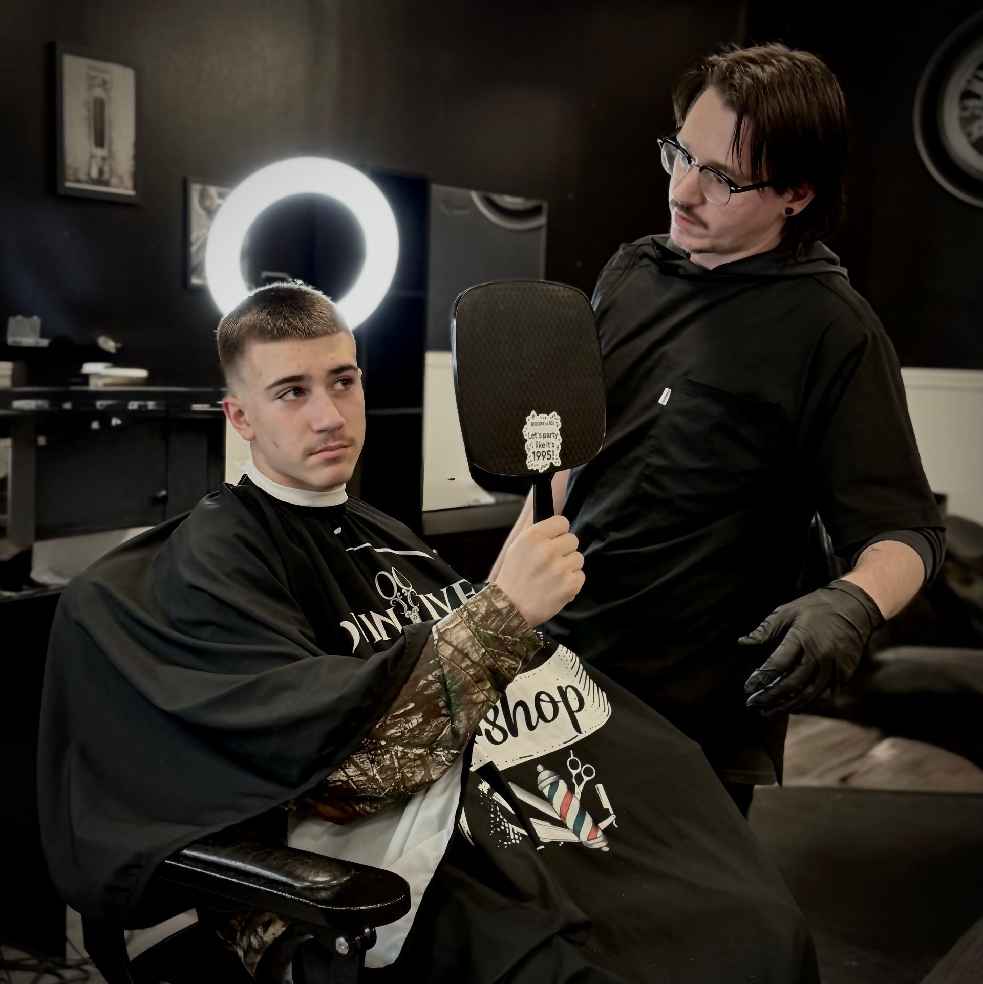 Barber showing a customer their haircut in a mirror. Both wear black clothing. Setting is a barber shop.