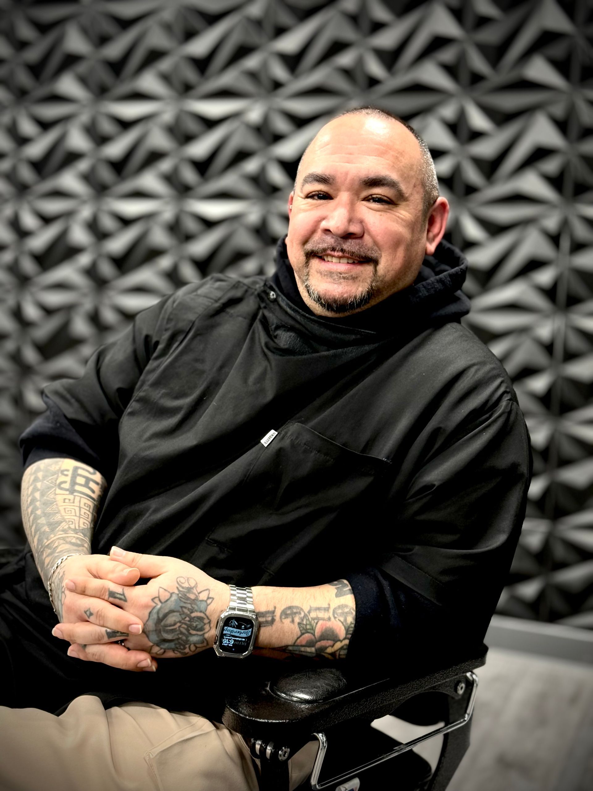Man in black smock smiles, seated. Tattoos on arms, against a textured gray wall.