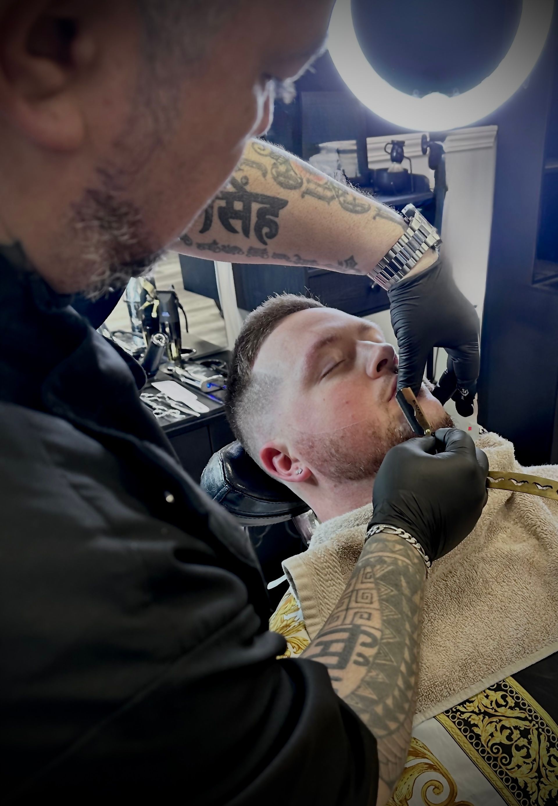 Barber shaving a man's face, close up. The barber wears black gloves and has tattooed arms.