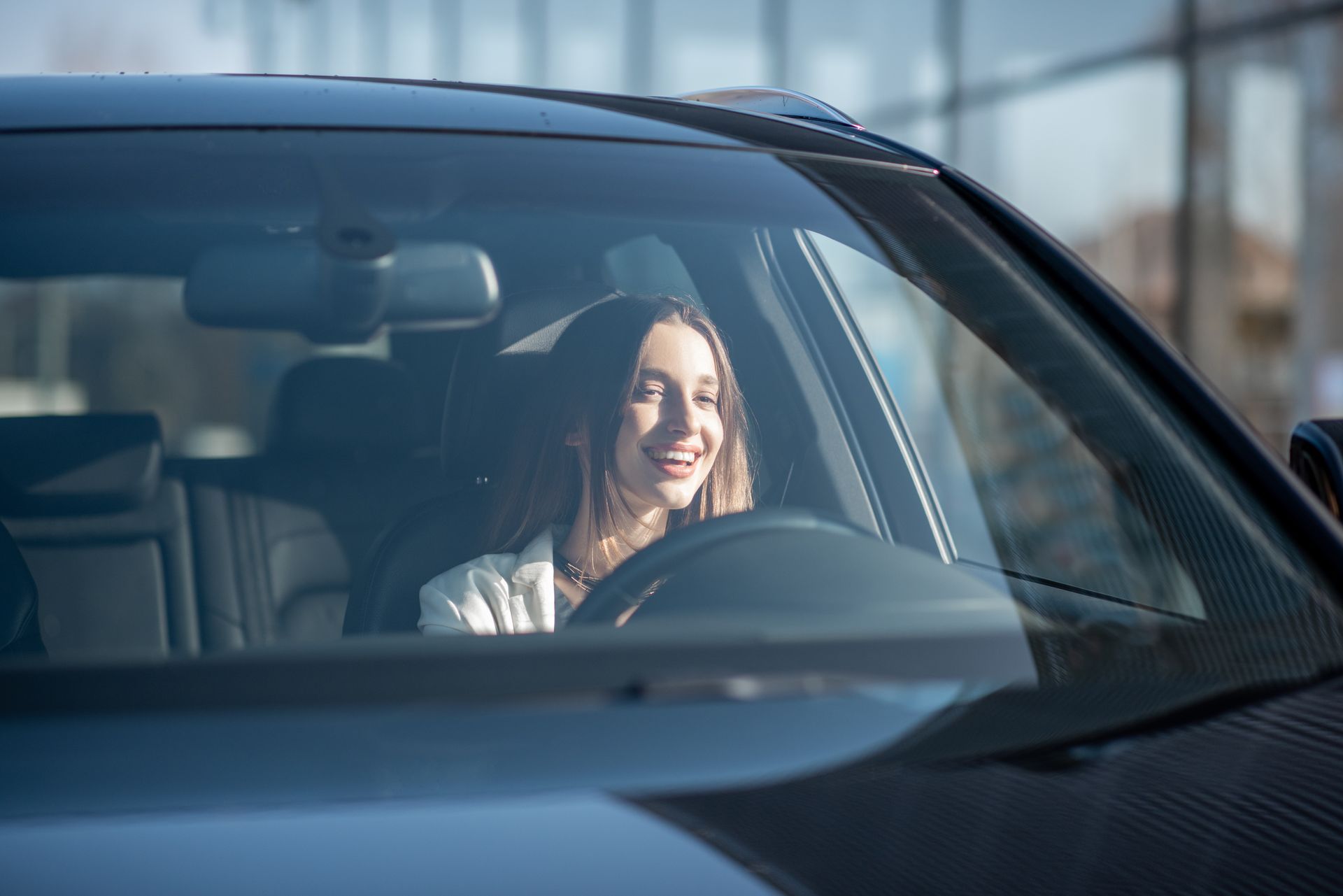 Woman smiling while driving a car. Her hands are on the steering wheel, and the interior is black.