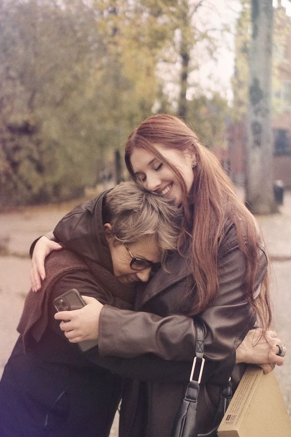 Two people hugging warmly outdoors beside a cardboard sign, with trees in the background.
