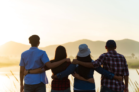 Four friends stand arm-in-arm facing a sunset over the water and distant hills.