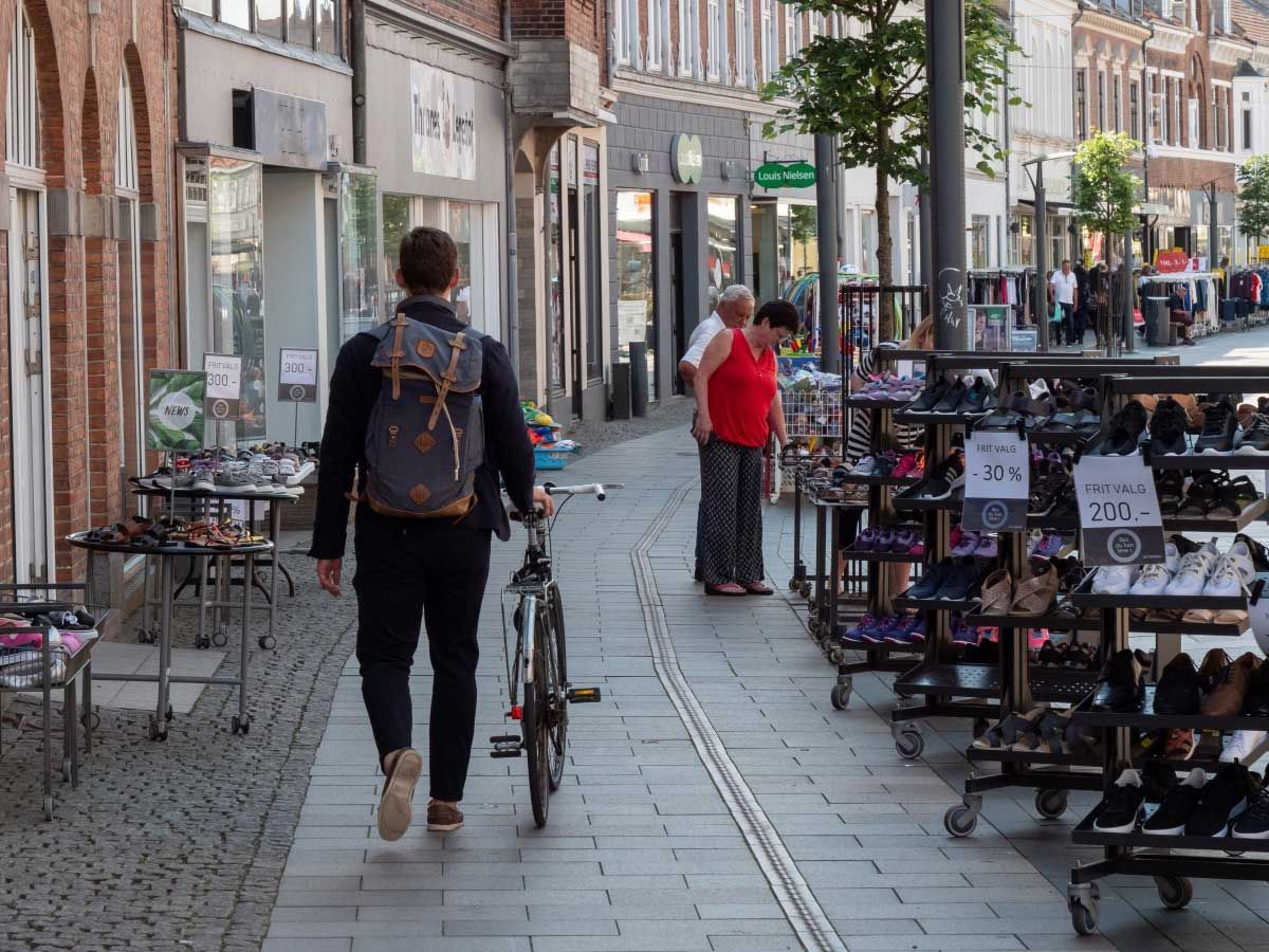 En mand med en rygsæk går ned ad et fortov med en cykel.