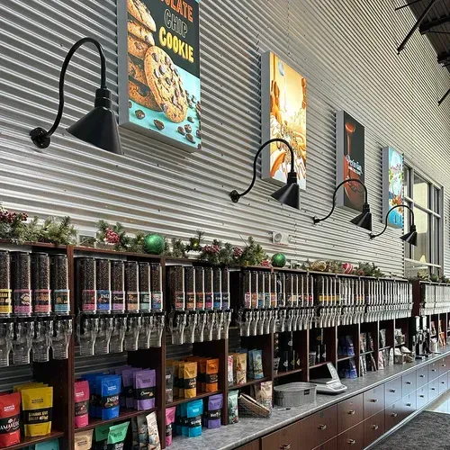 Coffee shop interior with coffee bean dispensers, posters, and decorative lights on a corrugated metal wall.