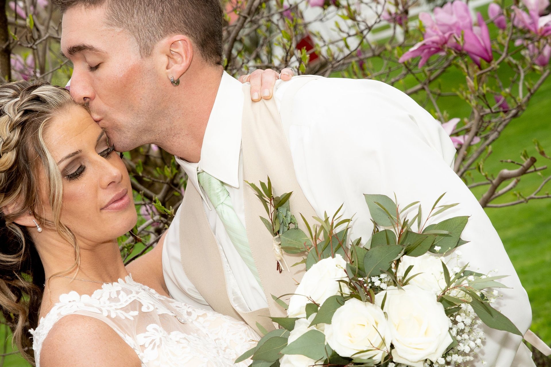 Groom kissing bride on forehead, holding bouquet. Outdoors with flowers in background, couple in wedding attire.