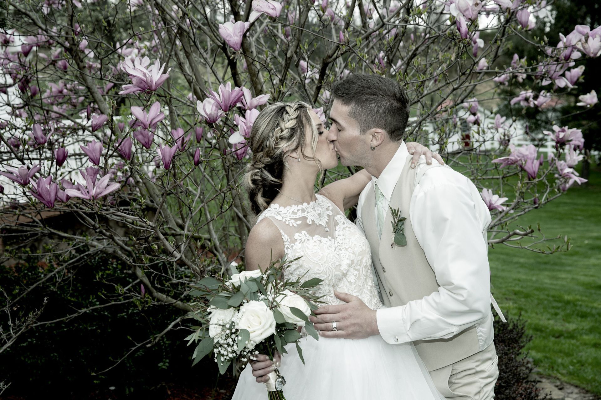 Bride and groom kissing, posed in front of a tree with pink flowers. She holds flowers. He wears a vest.