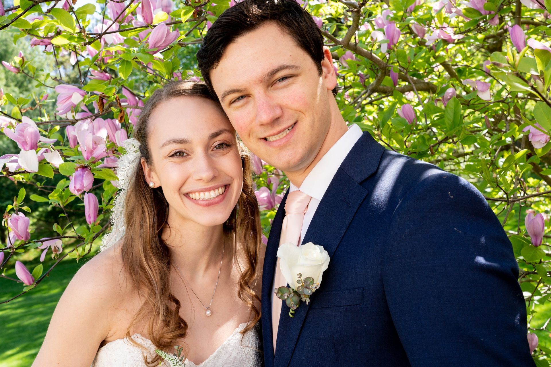 Bride and groom smiling under a blossoming pink tree. The man wears a blue suit, and the woman wears a white dress.