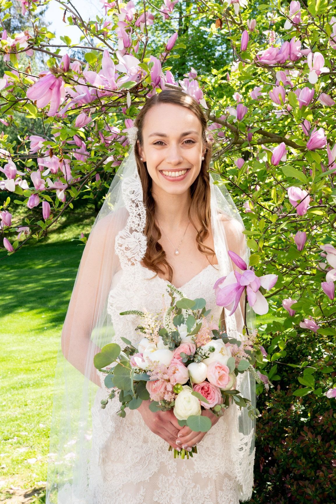 Bride in a white dress holding a bouquet, smiling in front of pink flowers and green foliage.