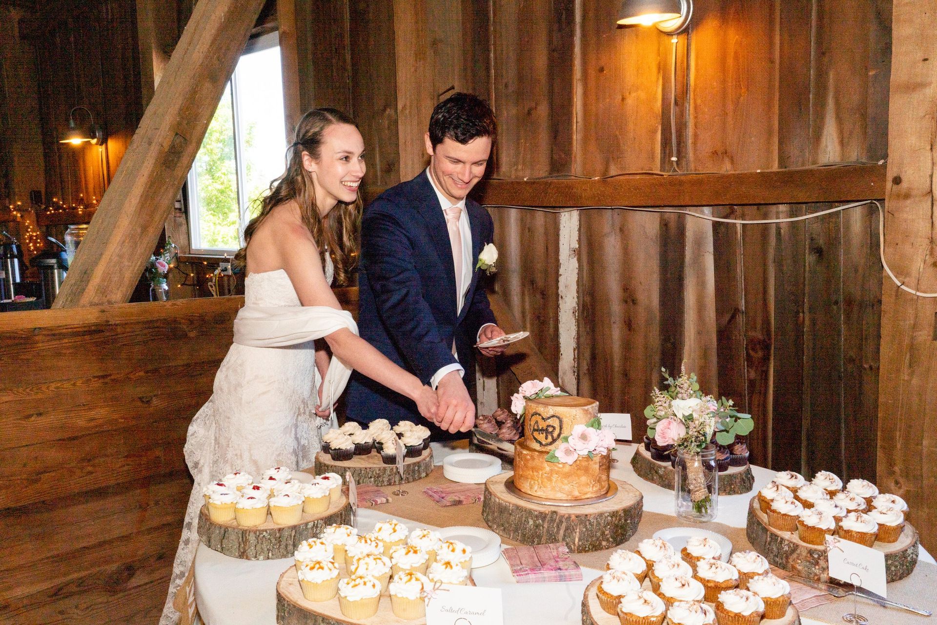 Couple cutting wedding cake in barn; cupcakes displayed.
