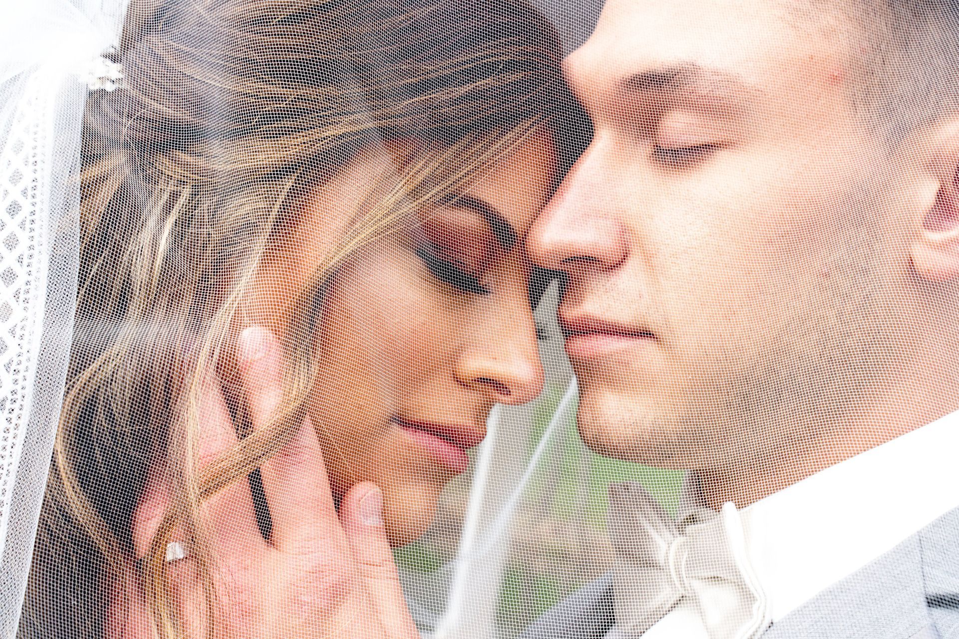 Bride and groom, faces close, under veil. Bride's hand on face, both with closed eyes.