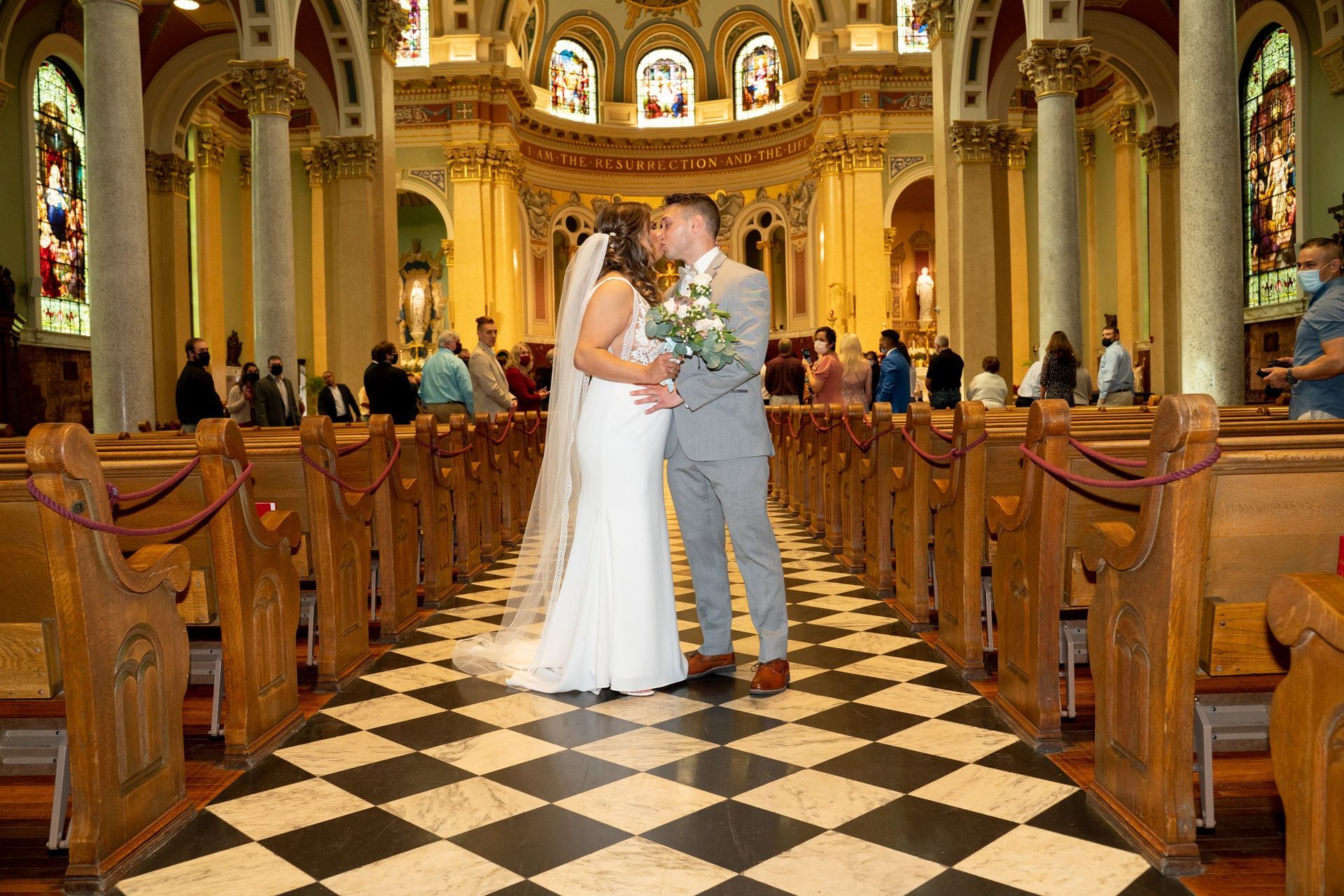 Bride and groom kiss at the altar in a church with checkerboard floor, wooden pews, and stained-glass windows.