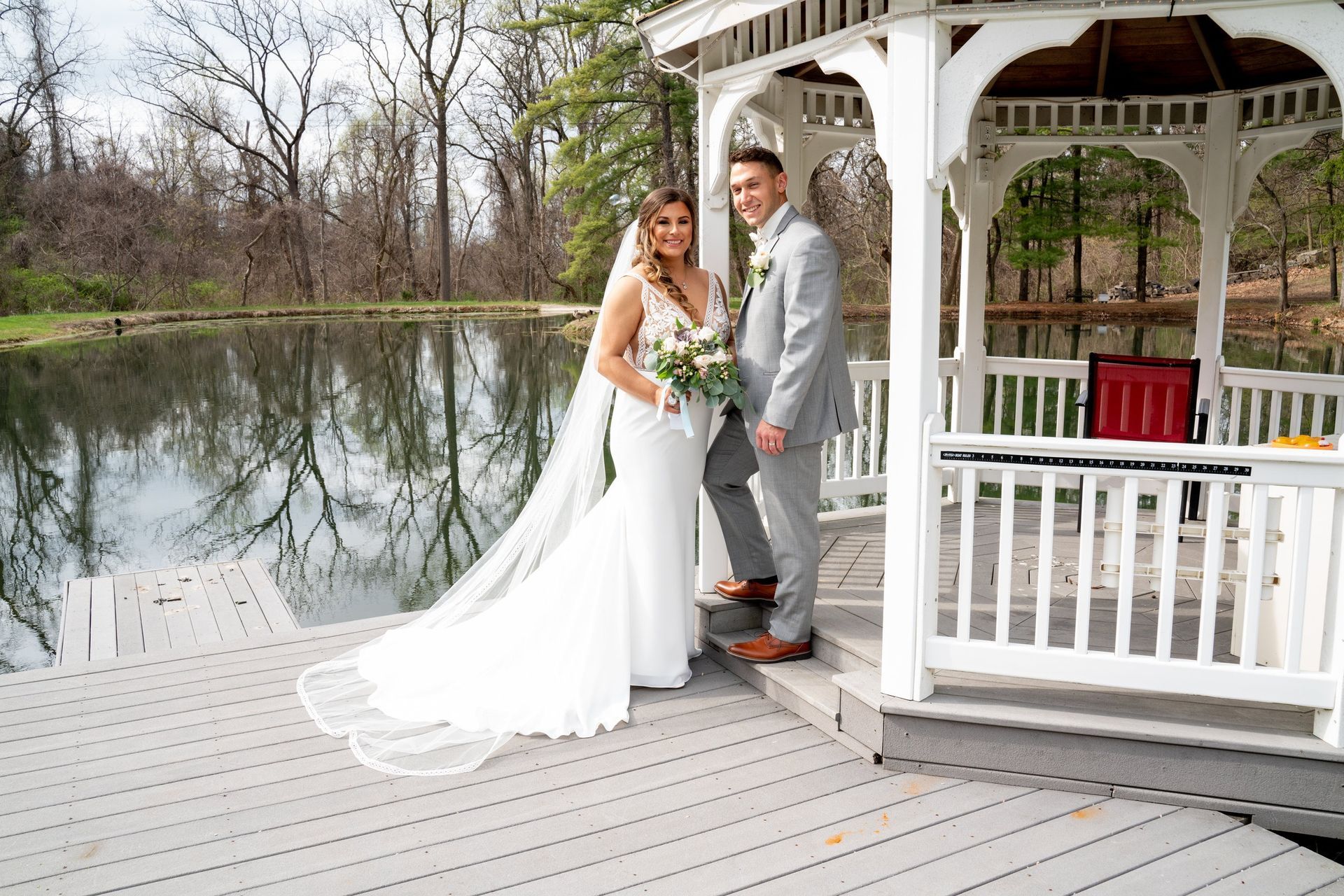 Newlyweds pose near a gazebo on a deck by a pond. The bride wears white, and the groom wears grey.