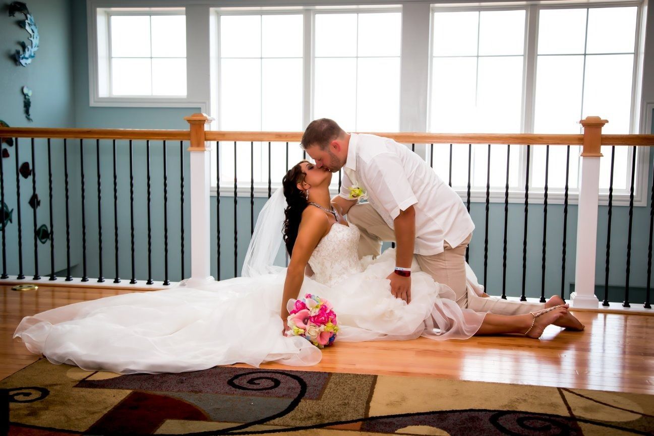 Bride in white dress lying on floor, groom kissing her, wedding setting with windows.