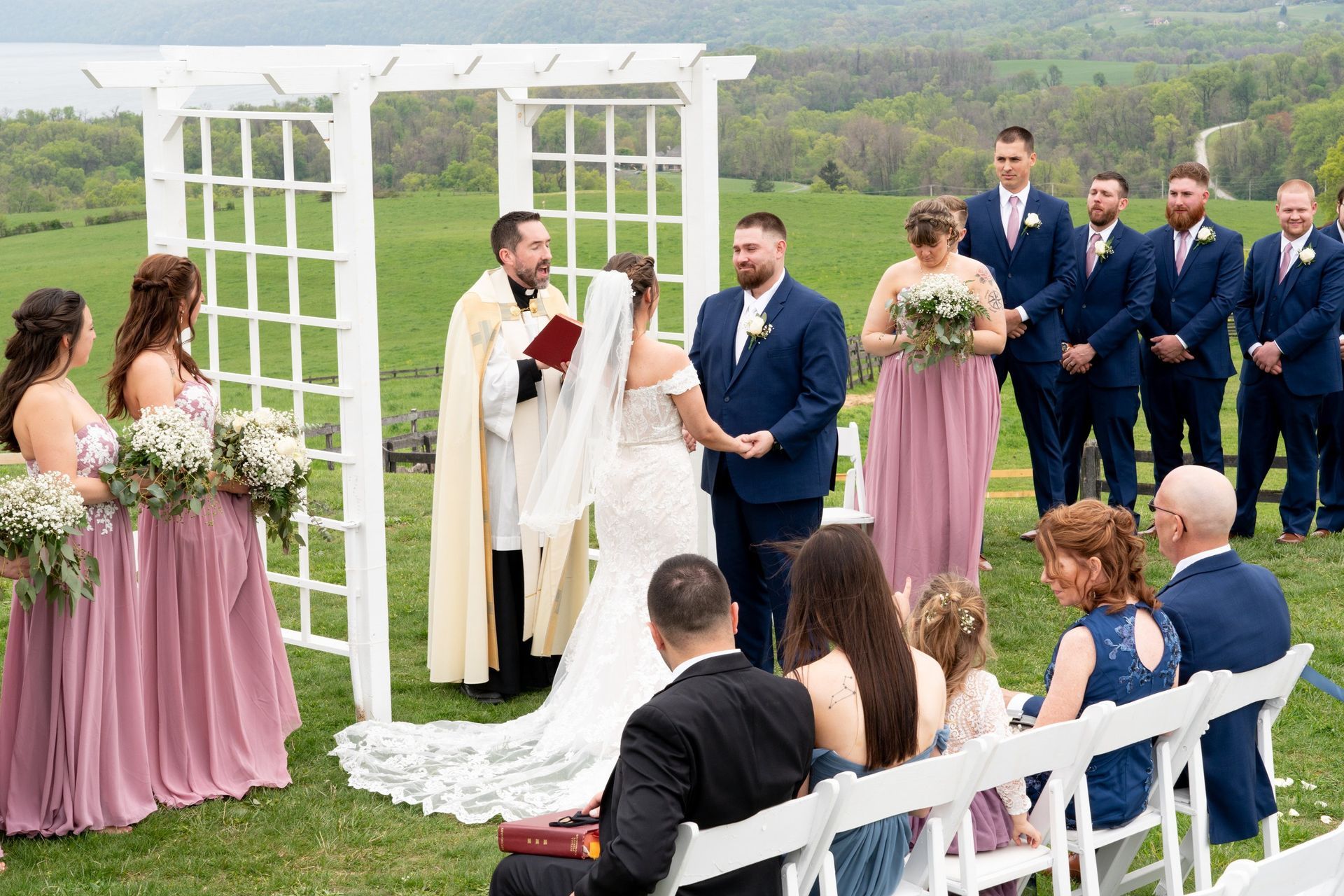 Wedding ceremony: Bride and groom with officiant under an arbor, bridal party, guests seated outdoors.