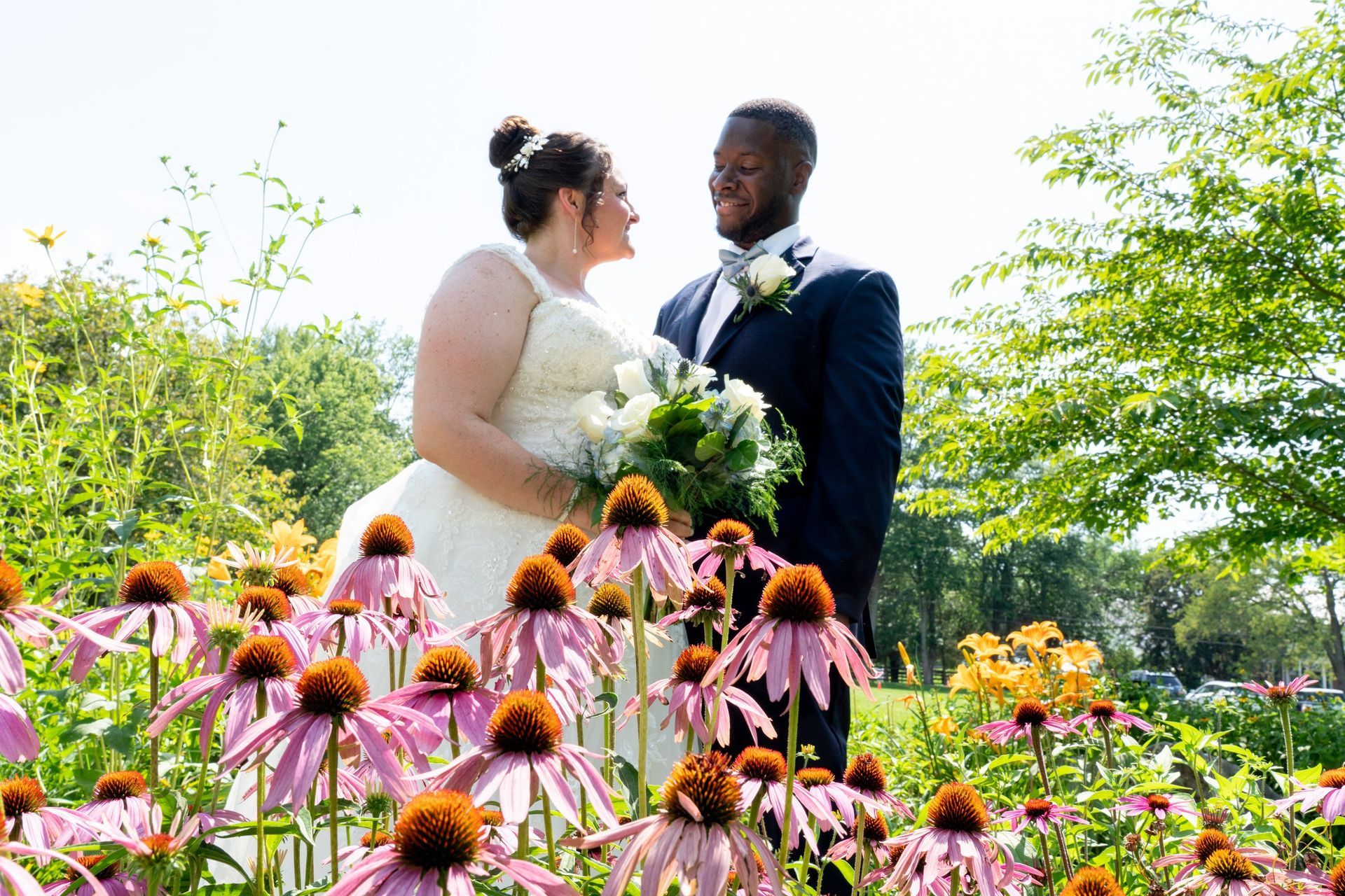 Bride and groom smiling at each other in a flower garden, she in a white dress, he in a blue suit.