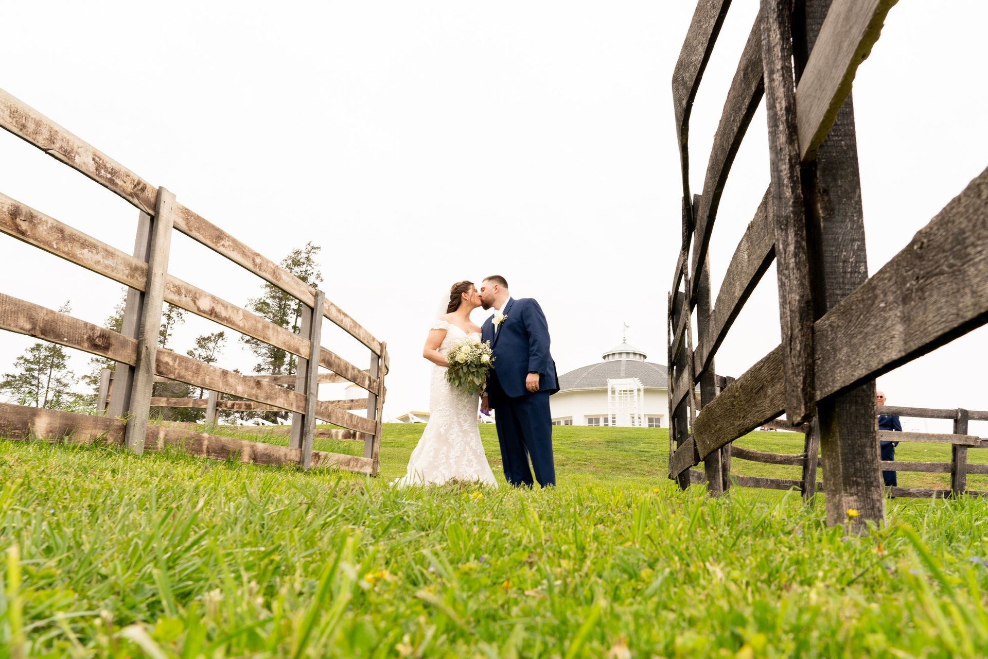 Bride and groom kissing in front of a wooden fence, with a white building in the background on a cloudy day.