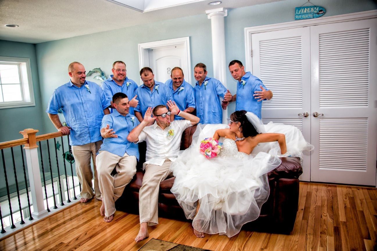 Groomsmen and bride playfully pose. Men in blue shirts and khaki pants, bride in gown on brown sofa.