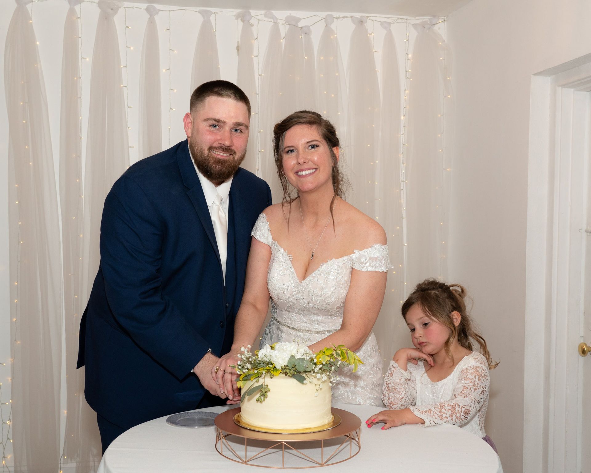 Couple cutting wedding cake with a flower girl; indoor setting, smiling, happy.