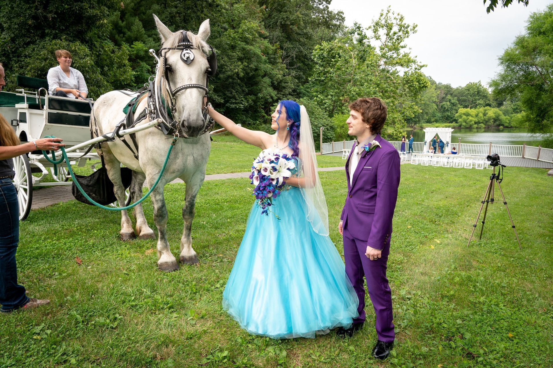Bride with blue hair and gown pets a white horse; groom in purple suit stands nearby. Outdoors.