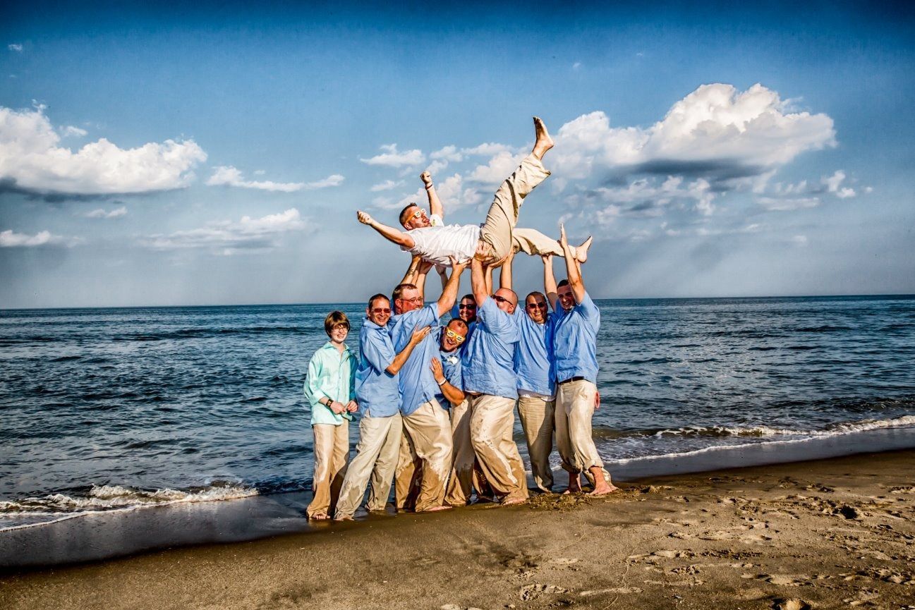 Men in blue shirts lift a man in white up in the air on a beach. Blue sky, ocean.