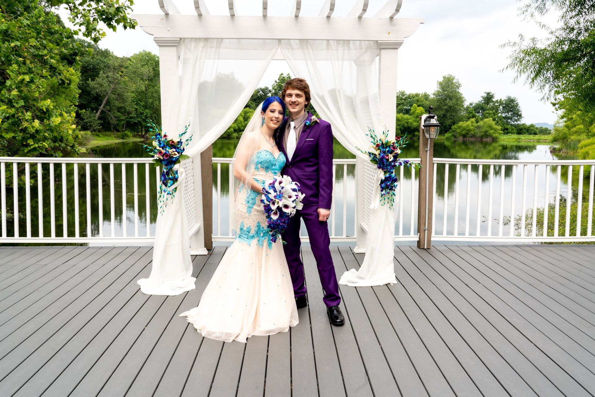 Couple in wedding attire pose on a deck before a decorated arbor overlooking a lake.