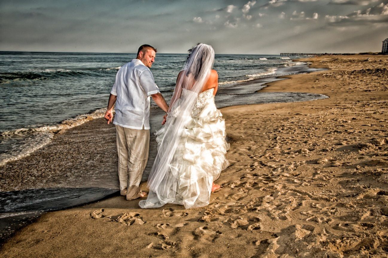 Newlyweds walk hand-in-hand on a beach at sunset, the bride in a white dress and veil.