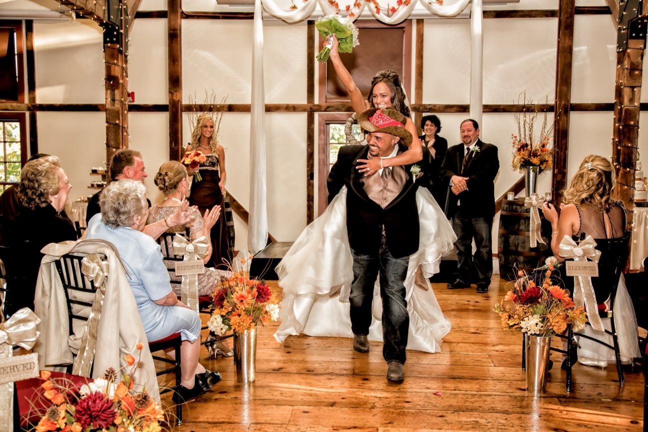 Bride on groom's shoulders, celebrating their wedding. Inside wooden building, guests applaud.