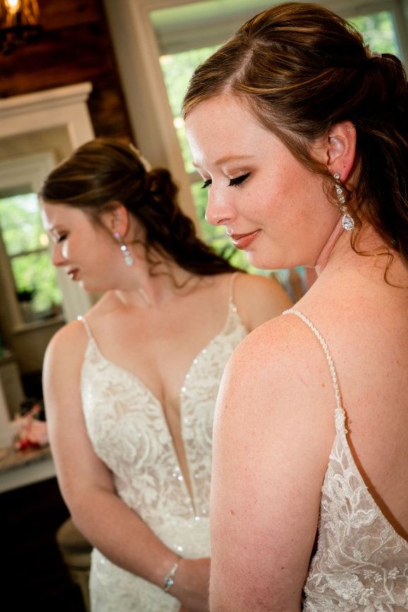 Woman in a white wedding dress looks in mirror, smiling. Indoors, fair skin, updo, with earrings.