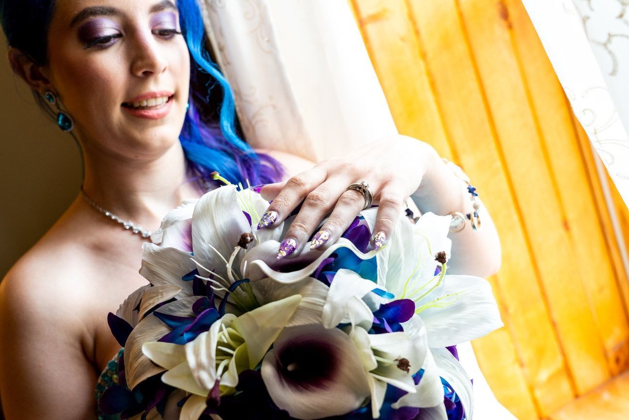 Bride with blue hair and purple eye makeup, holding white and purple bouquet, near a window.