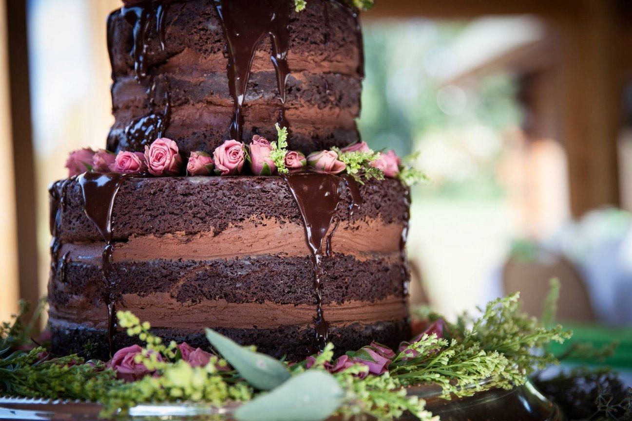 Three-tiered chocolate cake, dripping with glaze, decorated with pink roses and greenery; outdoors.