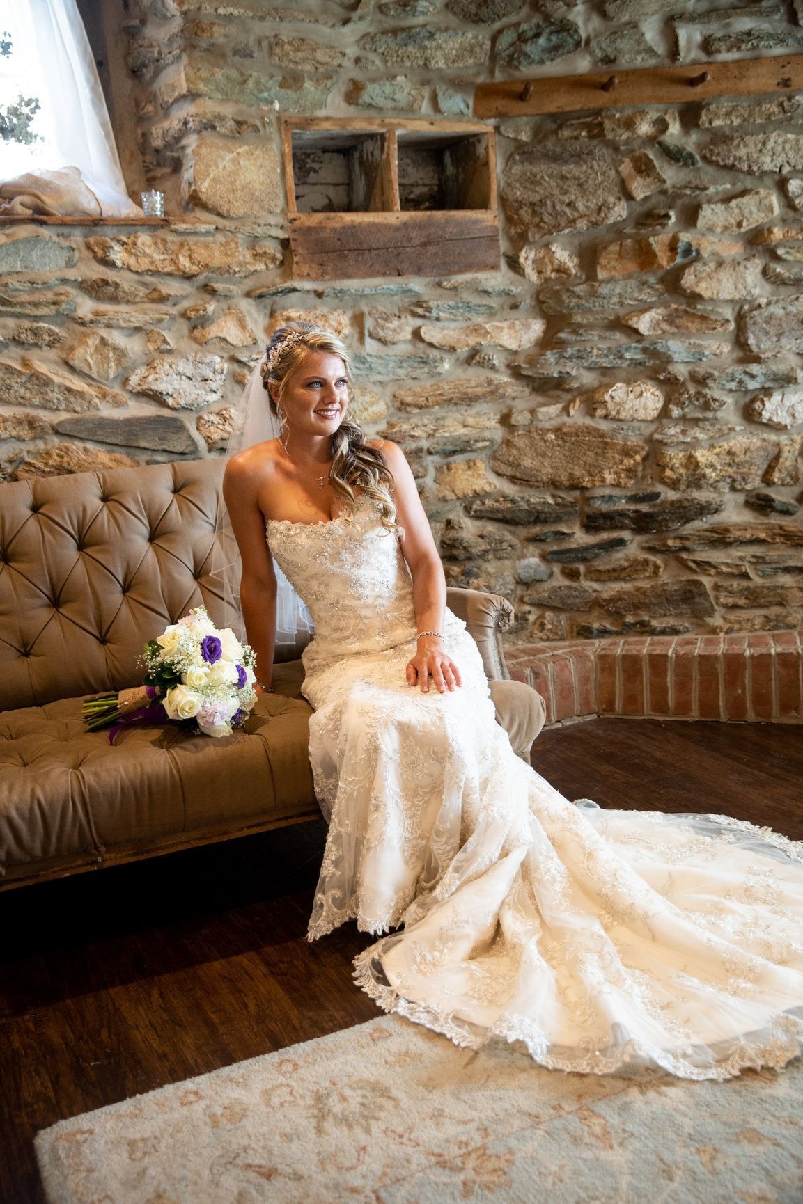 Bride in strapless lace gown smiles on brown tufted sofa, bouquet next to her. Rustic stone wall background.
